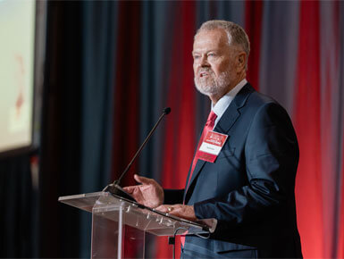 An older gentlman stands at a clear podium, giving a speech to the audience, with bright red and gray curtains behind him. He is wearing a dark suit.