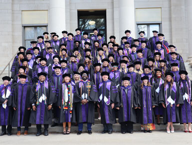The Knudson School of Law Class of 2026 stands on the steps of a building at USD, wearing their graduation regalia.