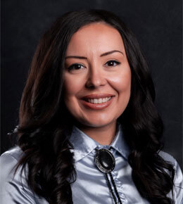 Cortni Bowman smiles at the camera for her professional photo, with dark hair, wearing a silver shirt and a black bolo tie against a dark background.
