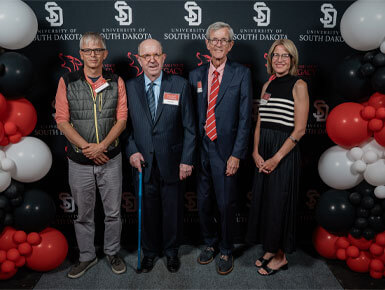Unleash Your Legacy recipients stand and smile at the camera, surrounded by red, white and black balloons, along with a dark backdrop with the white USD logo.