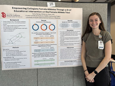 Carly Haring stands in front of a poster board, presenting her project, "Empowering Collegiate Female Athletes Through a Brief Educational Intervention on the Female Athlete Triad."