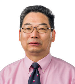 Xu Wang looks at the camera and smiles. He is wearing a pink button up shirt with a black and red tie, as well as glasses.
