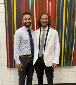 Sean McCann, stands with a friend, wearing a white lab coat and tie, standing against a backdrop of vertical colored stripes.