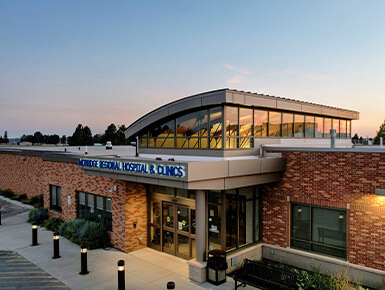 Exterior view of Mobridge Regional Hospital & Clinic at sunset, showing a brick building with large glass windows and a curved roofline.