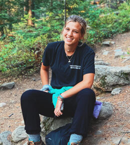 Beck Hofer sits on a rock on a hiking trail in a wooded area, smiling, wearing a dark t-shirt and dark pants.