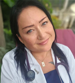 Amy Ortiz poses for the camera in a white lab coat and stethoscope around her neck, smiling with blue eyes, outdoors.