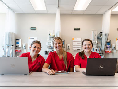 Three health care students wearing red scrubs sit at a desk with laptops in a clinical skills lab, smiling toward the camera, with hospital equipment and privacy curtains in the background.