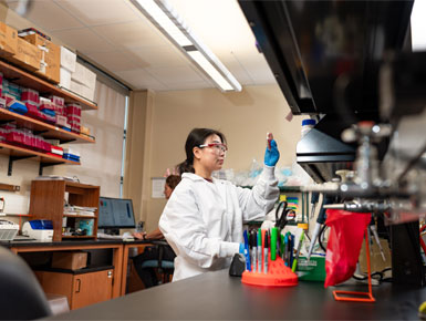 A USD researcher in a white lab coat, safety glasses and blue gloves holds up a small vial for inspection in a biomedical laboratory. She is surrounded by lab equipment, storage shelves with medical supplies, and a computer workstation in the background.