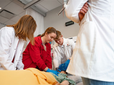 SSOM students in scrubs and lab coats practice a hands-on medical simulation, working together around a patient mannequin on a hospital bed in a clinical training room.