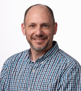 Victor Huber looks at the camera and smiles. He is wearing a black, blue, and white plaid button up shirt.