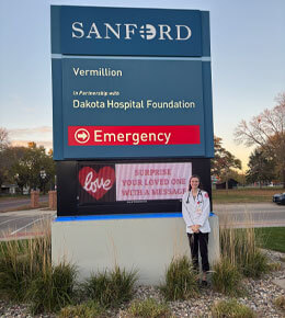 Carly Haring stands in front of a Sanford Health sign wearing her white coat. Haring is part of the USD FARM program.