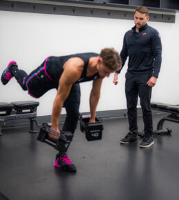 Hunter Glascock observes a player performing a single-leg dumbbell balance exercise in a gym.