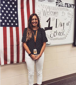 Paige Wright stands in her classroom in front of a white board. To her right is the American flag and to her left is the whiteboard that says, "Welcome to the first day of school."