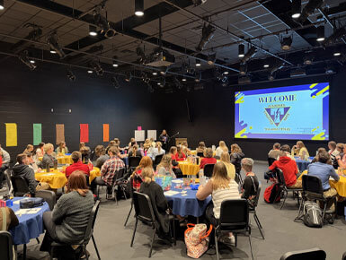 USD teacher candidates sit at round tables in a large room while a presentation is on the screen, showing a "Welcome" screen.