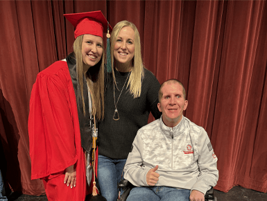 Kylie Briest stands in a red graduation cap and gown next to her parents, with her dad, who is sitting in a wheel-chair, giving a thumbs-up.