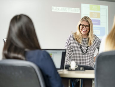 Emily Quinn stands in front of a screen, smiling at two students, during one of her marketing courses. The screen is displaying "Marketing" content.