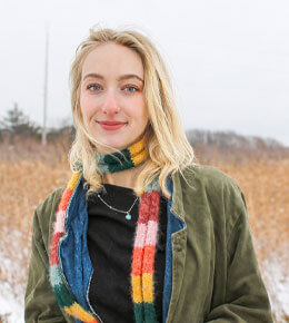 Bethany Larson stands in a wintry field, wearing a green jacket and colorful scarf. She is smiling at the camera.
