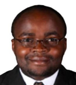 Etienne Gnimpieba sits for his professional photo. He is wearing a black suit and tie and smiling at the camera.