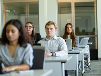 USD – Sioux Falls students sit in a classroom, looking forward toward their professor with their laptops sitting in front of them.