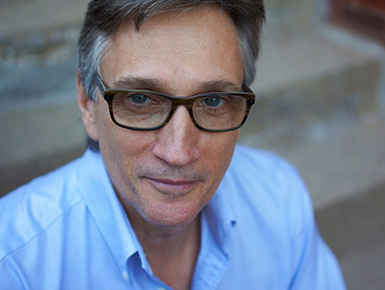 A headshot of a professional looking middle-aged man with glasses in a blue button down shirt in front of concrete steps