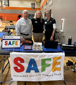 The GRACE organizers stand together behind a table in a gym during a fitness event. The table features a banner that reads, "SAFE: Sustained Athlete Fitness Exam."