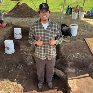 Angel Vazquez stands in a dug out hole surrounded by dirt and anthropology resources.
