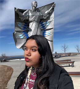 Tyler LaMere stands in front of a Native American statue, wearing a black coat. The statue has outstreatched arms, with a blanket behind it, and is silver and blue.