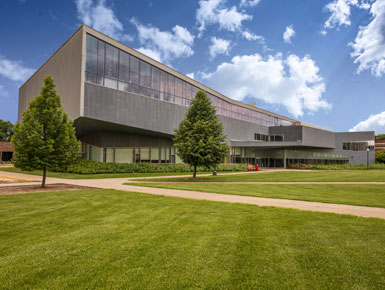 Beacom Hall, home of the Beacom School of Business, stands on the University of South Dakota campus, surrounded by green grass and trees beneath a sunny blue sky with a few clouds.