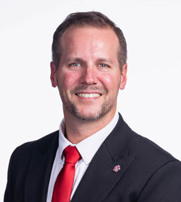 USD Athletic Director Jon Schemmel poses for his professional photo. He smiling and wearing a black suit jacket, red tie and white shirt, along with a USD pin.