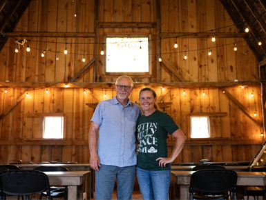 Jeff and Nancy Kirstein stand together in the loft of a barn. There are string lights hanging on the back wall and tables and chairs spread around the space behind them.
