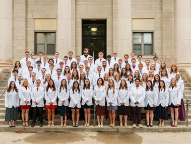 The USD Sanford School of Medicine Class of 2030 stands on the steps of a campus building. In the background are grand pillars and windows.