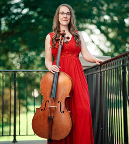 Sonja Kraus stands on an outdoor patio, with her left hand resting on a black fence, with her right hand clutching her cello. She is wearing a bright red dress and the background is filled with green trees.