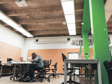 A student studies at a desk in a quiet, industrial classroom, surrounded by tables and chairs, with vivid green columns and bright fluorescent lights.