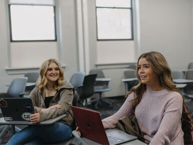 Two female USD students sit in a classroom at desks. They each have a laptop in front of them, are smiling and are looking towards someone who isn't in the camera.