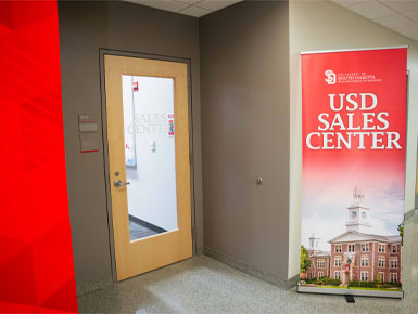 A hallway is lined with gray walls and carpet, a wood-framed glass door to the Beacom School of Business Sales Center and a red USD banner featuring campus imagery.