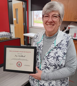 Joy Molland stands in an office setting, holding her CSA award certificate and smiling at the camera.