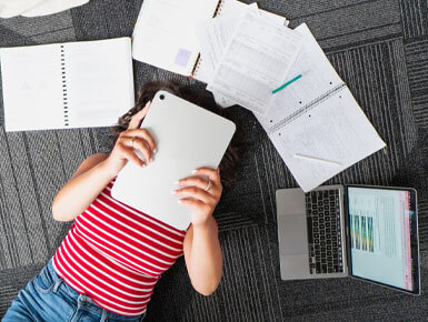 A student studies on the floor with her iPad, laptops and notes surrounding her.