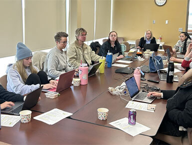 Cheryl Hartman sits around a conference table with her students during a class. Some are smiling at the camera while others are looking at their laptops and notes.