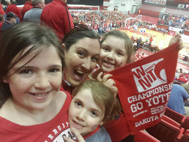Kari Oyen and her children pose for a photo in a sports arena. They are dressed in Coyote gear and holding a "Champions! Go Yotes!" flag.