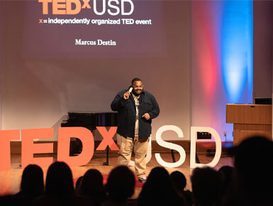 Marcus Destin stands on a stage while presenting his TedTalk. He is speaking to a crowd with a screen behind him.
