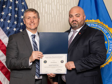 Bryant Jackson stands with a colleague, who is awarding him a certificate. The State of South Dakota flag and the U.S. flag are behind them.